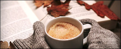sweater covered hands clasp a mug of hot cider in front of a book with fall leaves scattered on top